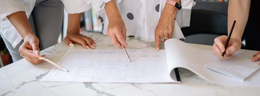 A picture of two people's hands pointing down on construction plans and another set of hands writing on a note pad.